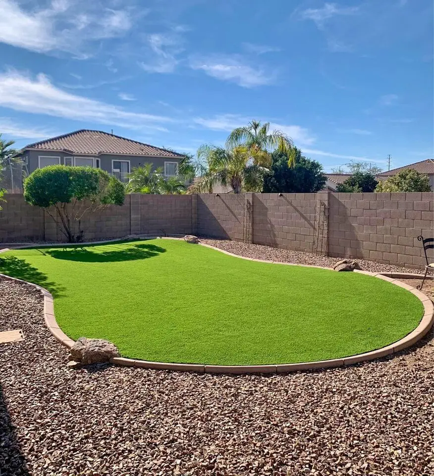 A long driveway with a freshly washed and finished concrete surface, courtesy of top-tier Concrete Contractors Miami, leads to a residential garage. The driveway is bordered by red bricks and surrounded by garden beds and neatly trimmed bushes. A garden hose lays on the left, with wrapped objects on the right side.